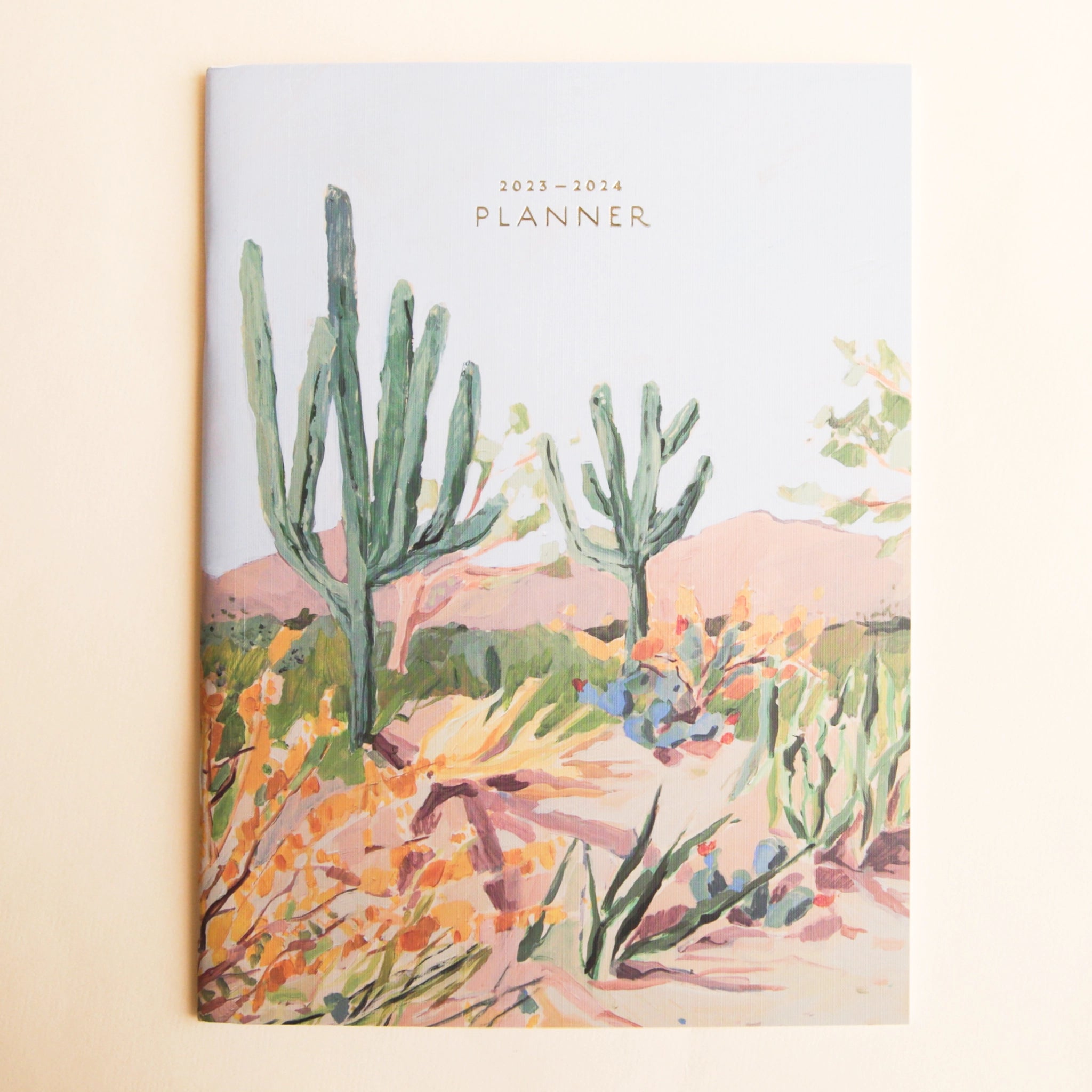 On a tan background is a cardstock planner with a beautiful desert scape illustration on the front along with small text at the top that reads, &quot;2023-2024 Planner&quot;.