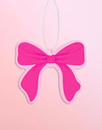 A hot pink bow with light pink trim air freshener is shown against a light pink to peach gradient background.