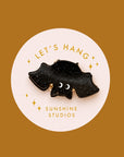A black speckled bat shaped mini hair clip on a round piece of packaging that reads, 'Let's Hang Sunshine Studios'.