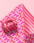 A pink and red piece of wrapping paper with a reversible pattern. One side features a bow pattern and the other, a pink stripe. Box and bow not included with purchase.