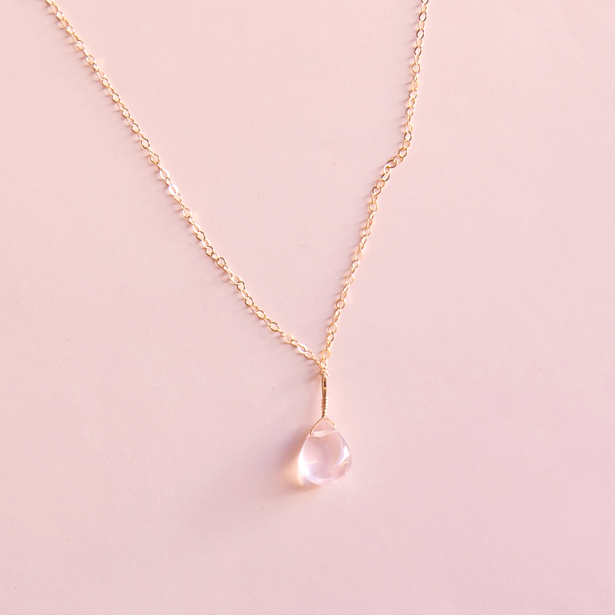 On a neutral background is a gold chain necklace with a rose quartz stone in the center.