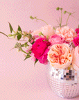 On a light pink background is a silver hanging planter with a pink flower arrangement in it.