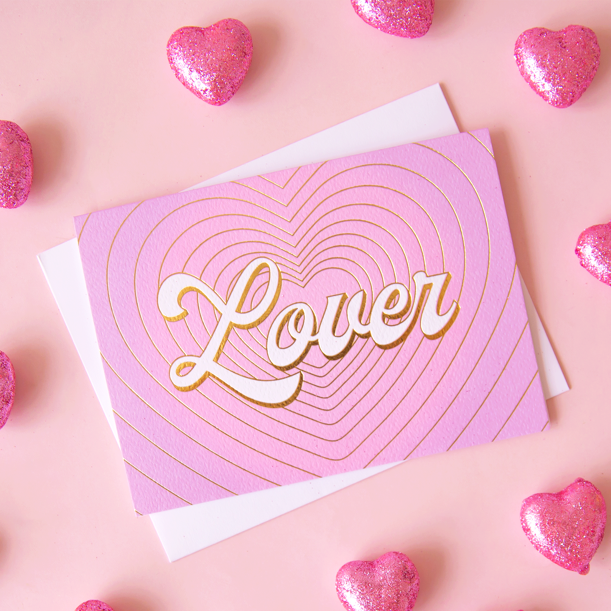 On a pink background is a pink and purple gradient card with gold foiled heart shaped outlines and ivory text that reads, &quot;Lover&quot;. 