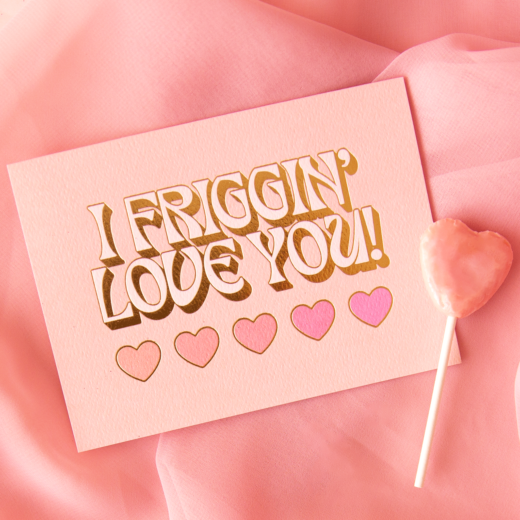 On a pink background is a light pink card with text that reads, &quot;I Friggin&#39; Love You!&quot; with five hearts underneath and staged next to a heart shaped sucker that is not included with purchase. 