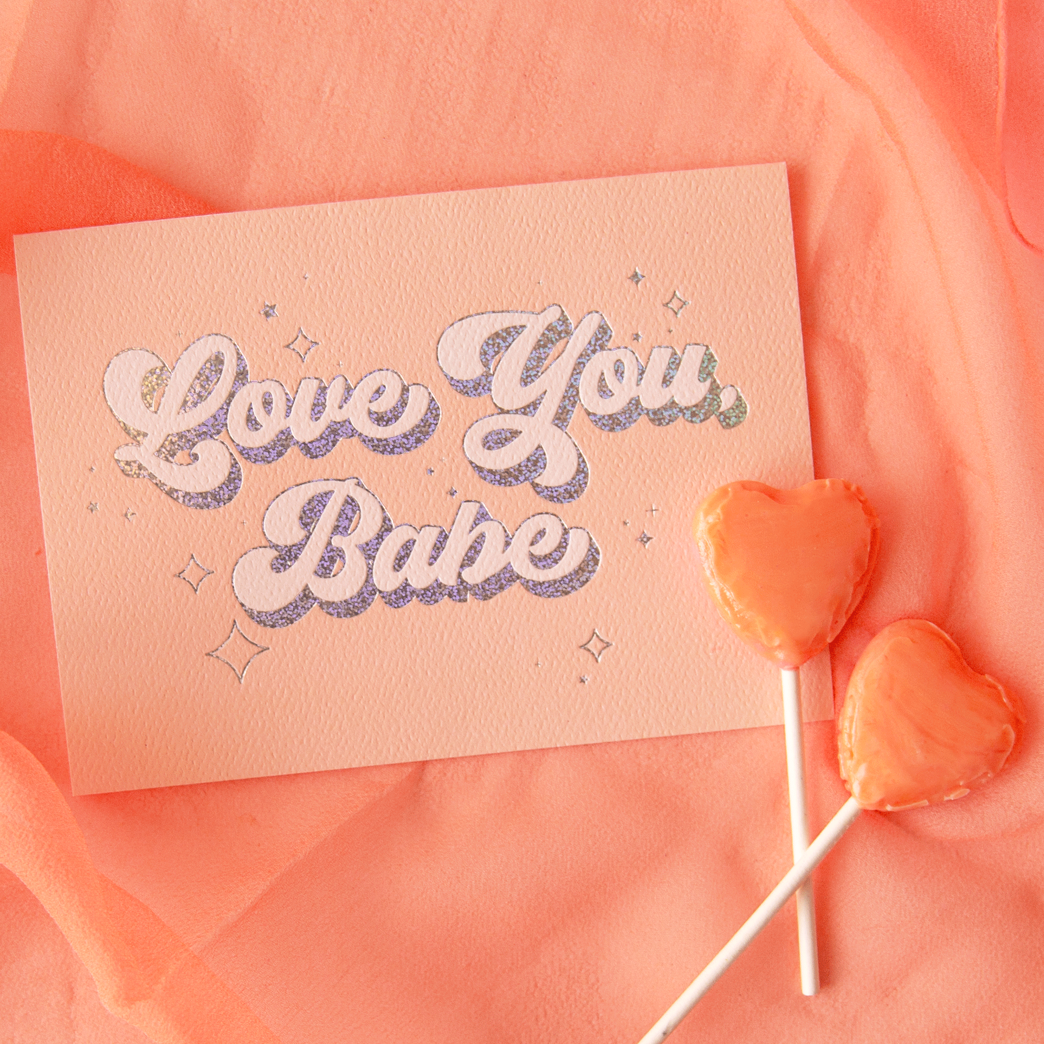 On a peach background is a peachy greeting card with &quot;Love You Babe&quot; text in the center that is outlined with holographic foil detailing. Also included is a white envelope.