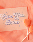 On a peach background is a peachy greeting card with "Love You Babe" text in the center that is outlined with holographic foil detailing. Also included is a white envelope.