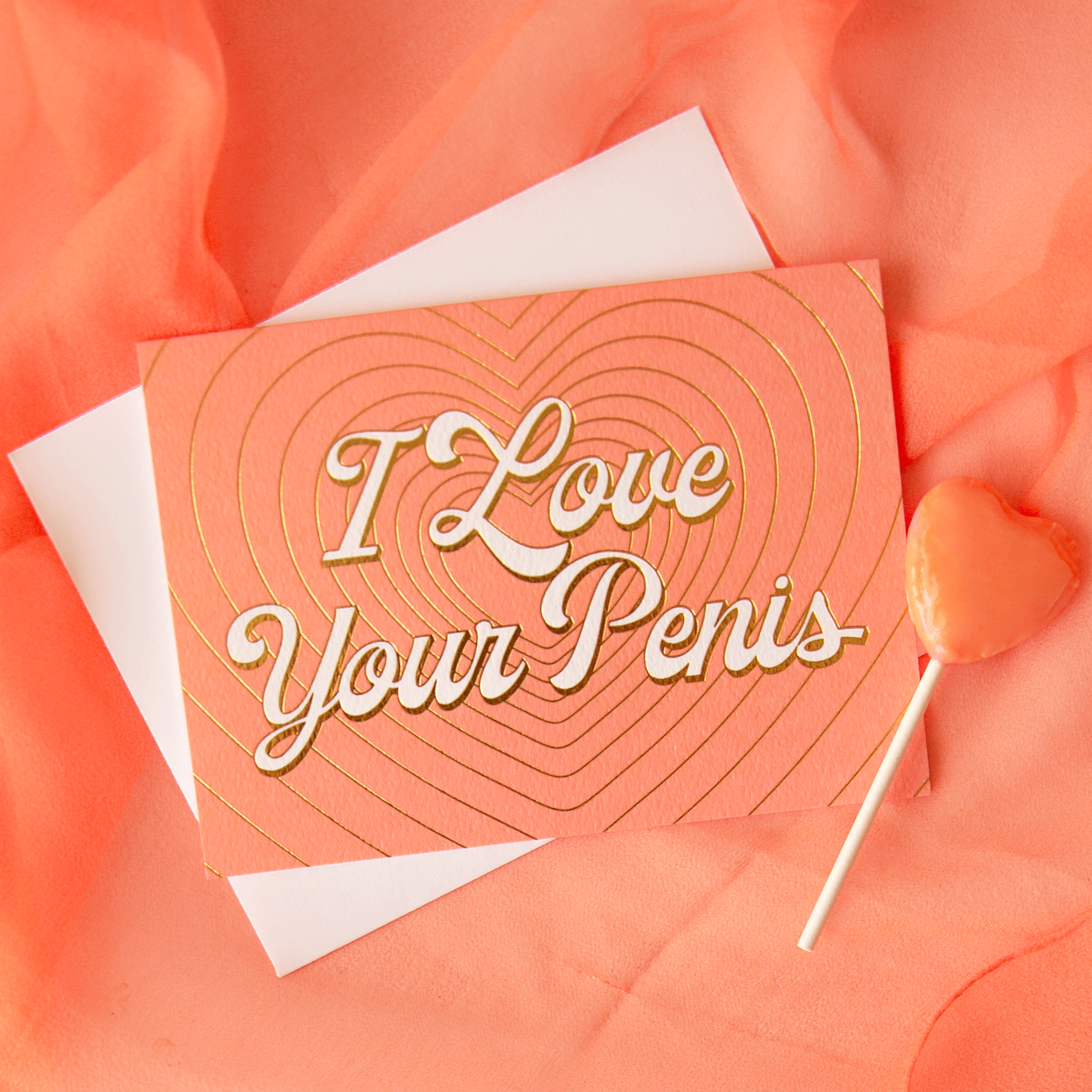 On a peachy background is a peachy colored card with layered gold foiled hearts and ivory text that reads, &quot;I Love Your Penis&quot; along with a white envelope.