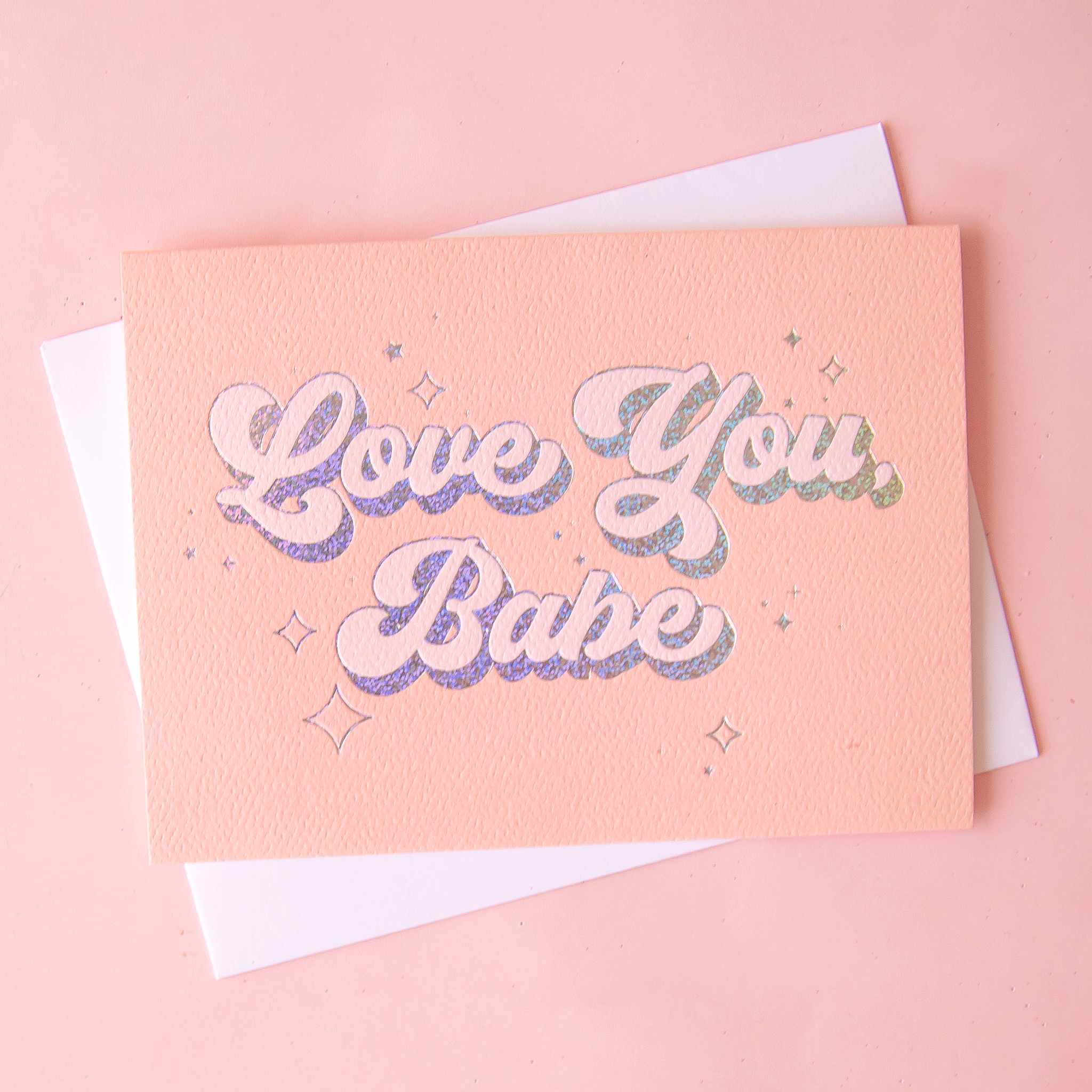 On a pink background is a peachy greeting card with "Love You Babe" text in the center that is outlined with holographic foil detailing. Also included is a white envelope.