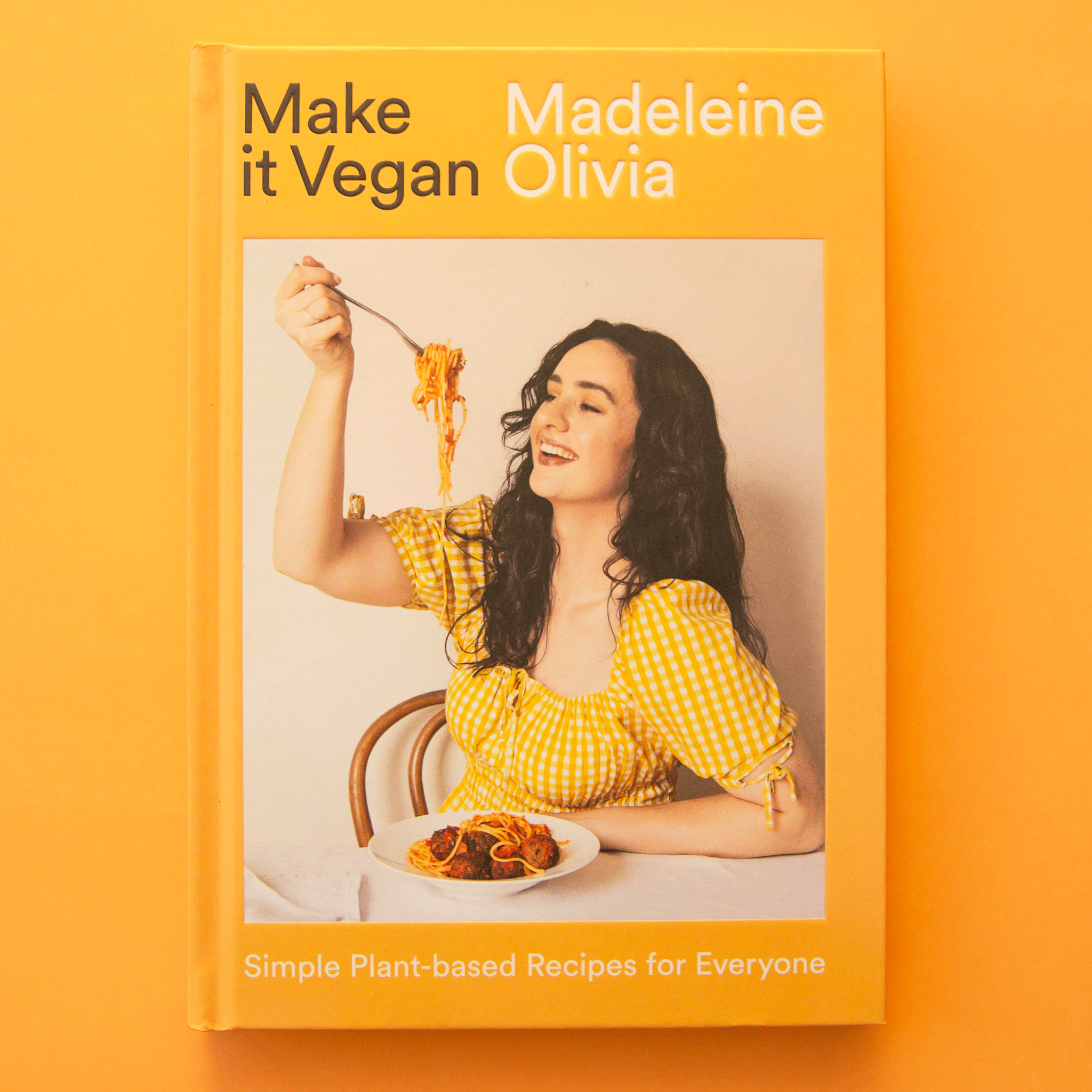 On a yellow background is a yellow cookbook cover with a model holding up a fork full of spaghetti along with black and white text above and below the image that reads, "Make it Vegan Simple Plant-based Recipes for Everyone". 