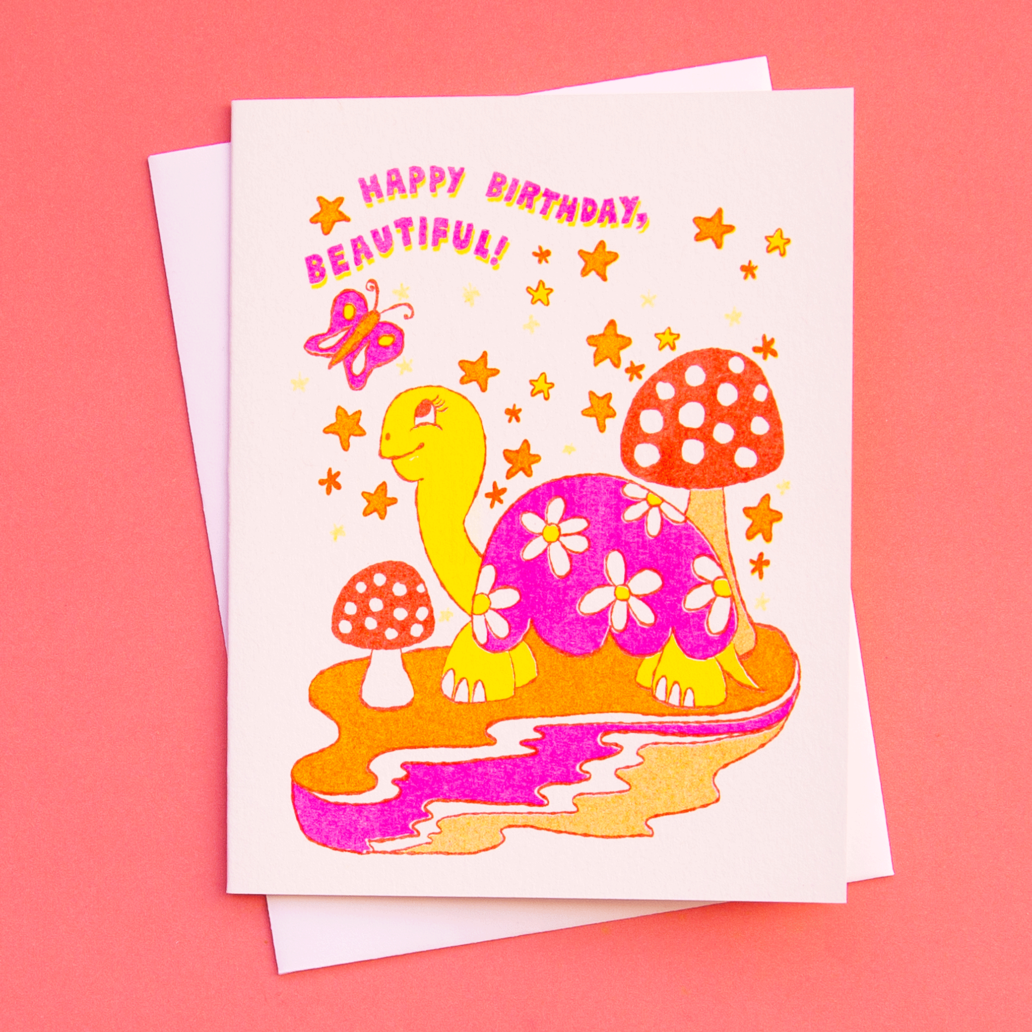 On a coral background is a white card with a colorful, red, pink and orange turtle illustration surrounded by mushroom, stars and a butterfly along with text that reads, "Happy Birthday Beautiful!".