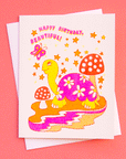 On a coral background is a white card with a colorful, red, pink and orange turtle illustration surrounded by mushroom, stars and a butterfly along with text that reads, "Happy Birthday Beautiful!".