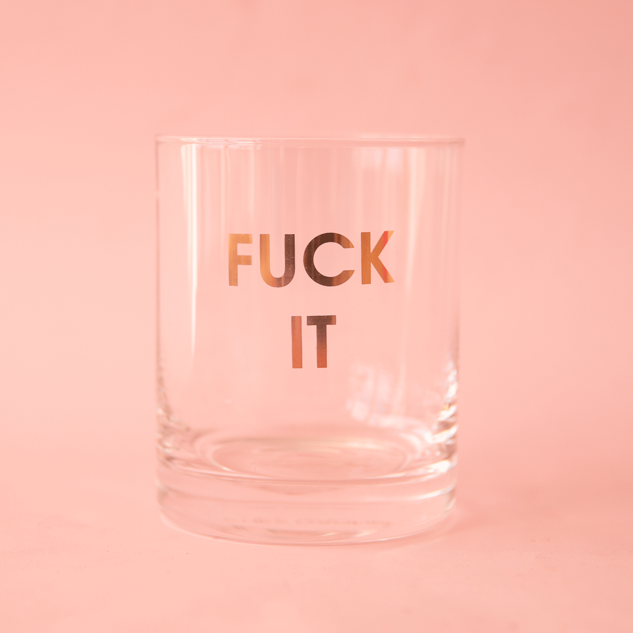 On a pink background is a clear glass mug with gold text across the front that reads, "Fuck It".