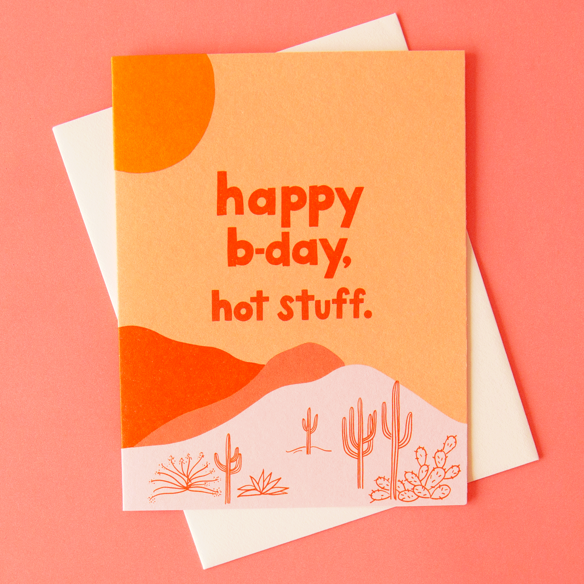 A salmon pink greeting card with a desert scape graphic featuring cactus and aloe illustrations along with a red and pink mountain range and a mustard yellow sun in the top left corner. The text on the front reads, "happy b-day hot stuff." in red font.
