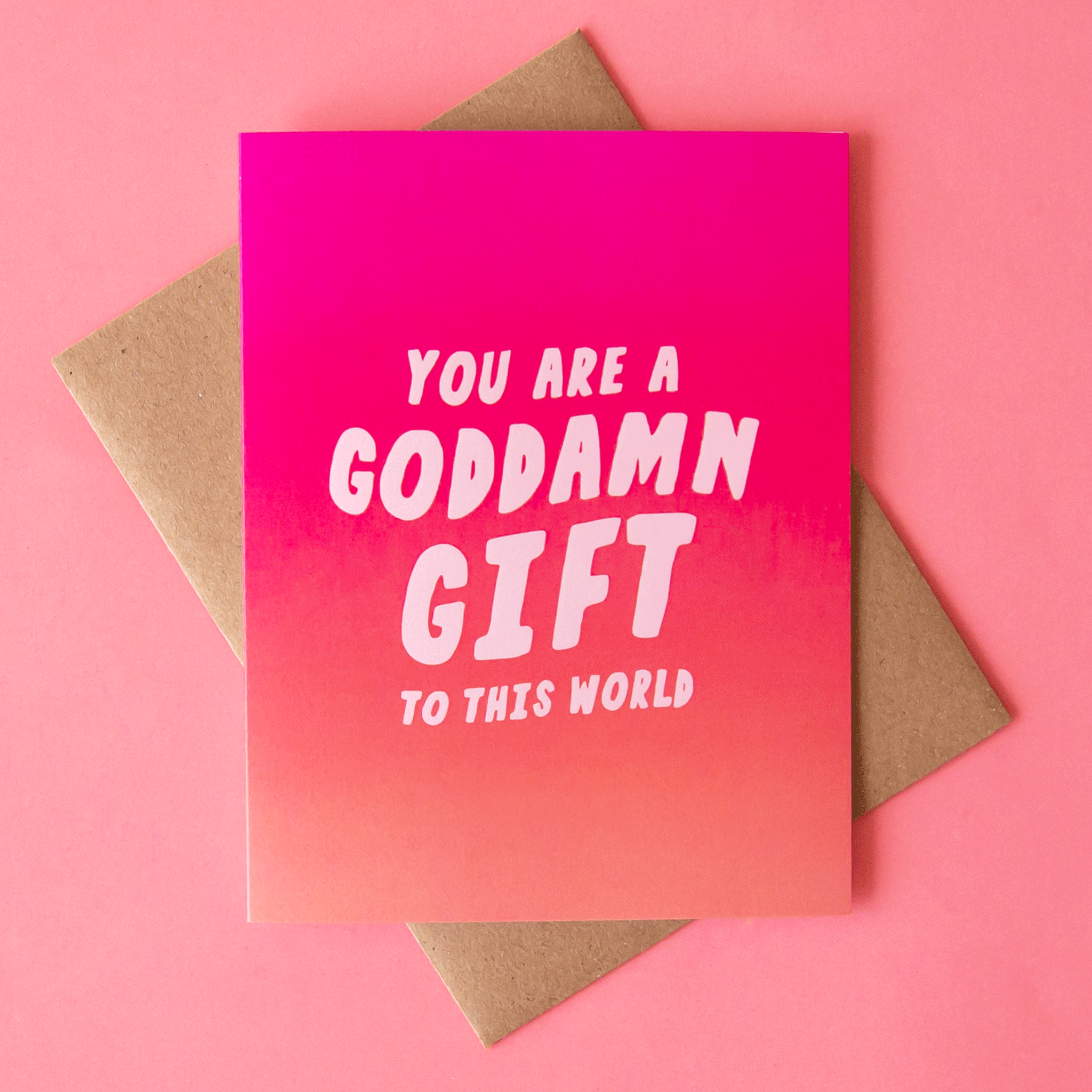 This card reads 'You Are a Goddamn Gift to This World' with a background featuring an ombre of rosie tones. This card is accompanied by a kraft brown envelope.