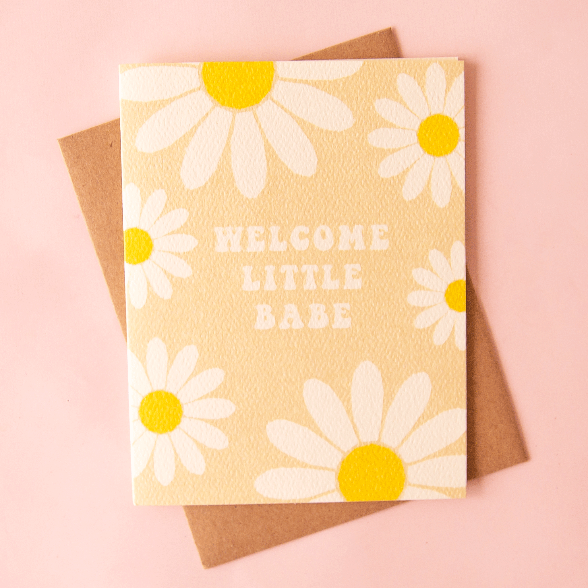 A tan card with white and yellow daisies all over the edges along with groovy text in the center that reads, "Welcome Little Babe" along with a Kraft brown envelope.