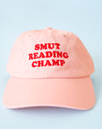 A pink baseball hat with red text that reads, 'Smut Reading Champ'.