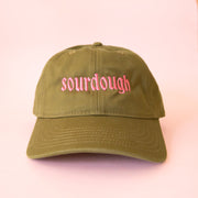 An army green baseball cap with pink text that reads, 'sourdough'.