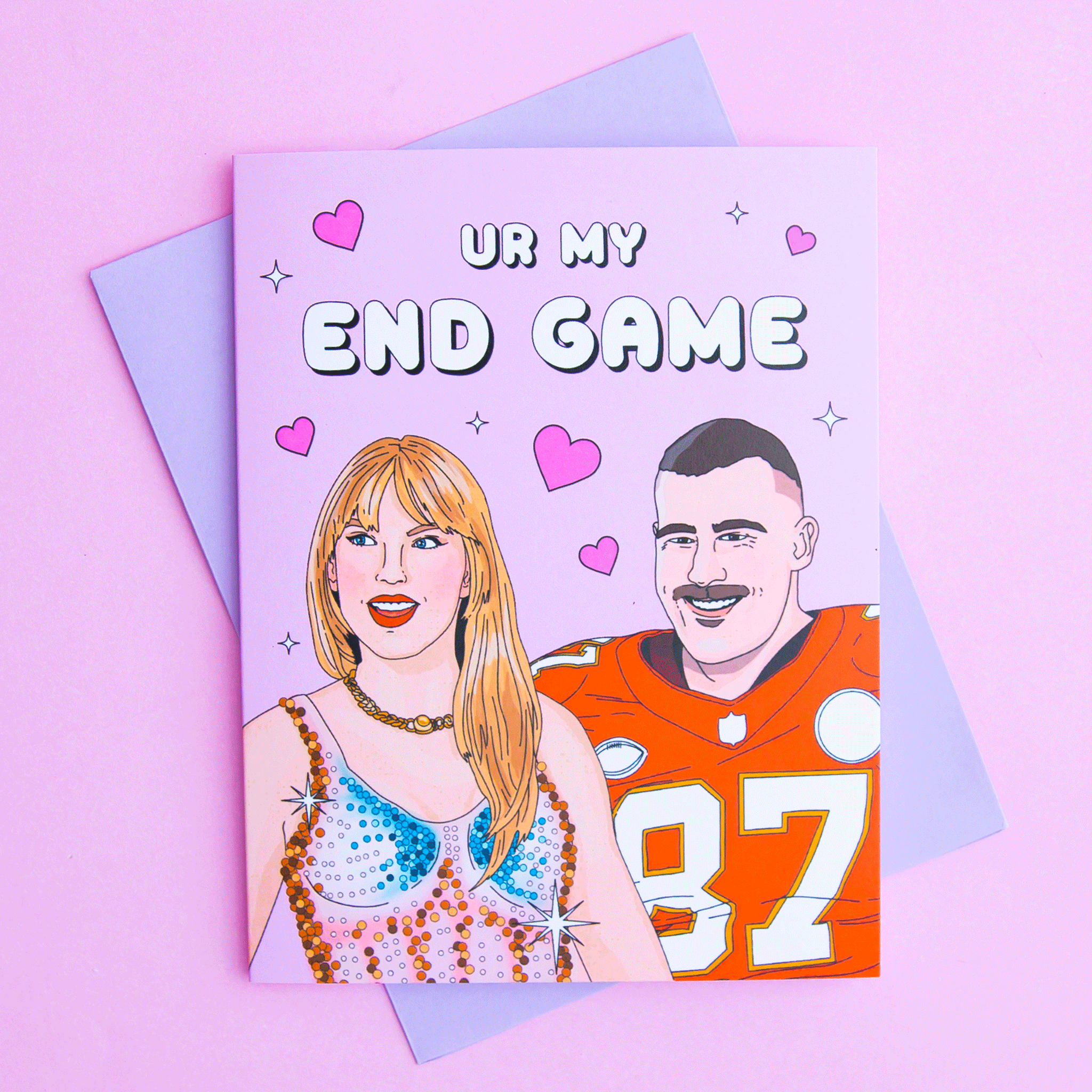 On a pink background is a pink card with an illustration that looks like Taylor Swift and Travis Kelce along with text above them that reads, &quot;Ur My End Game&quot;. The card comes with a light purple envelope.