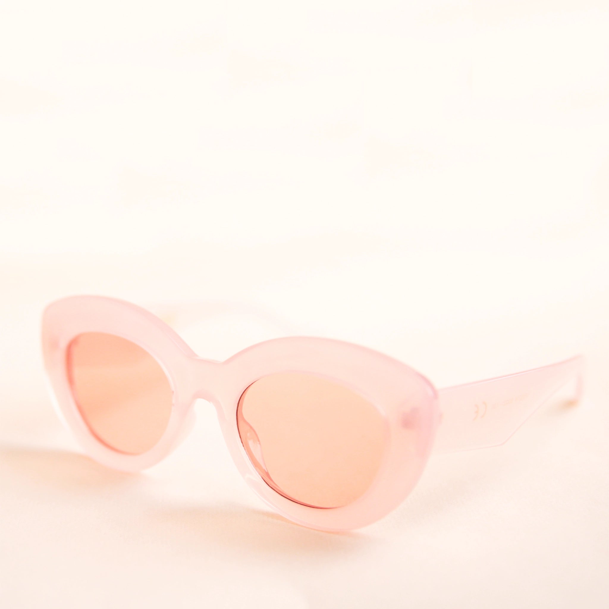 On a neutral background is the Gemma Sunglasses in a light pink shade. They have a rounded shape with a slight cateye flair at the corners. The frame material is a durable plastic and the lenses are a similar tone.