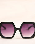 On a neutral background is a pair of hexagon shaped black sunglasses with purple lenses.