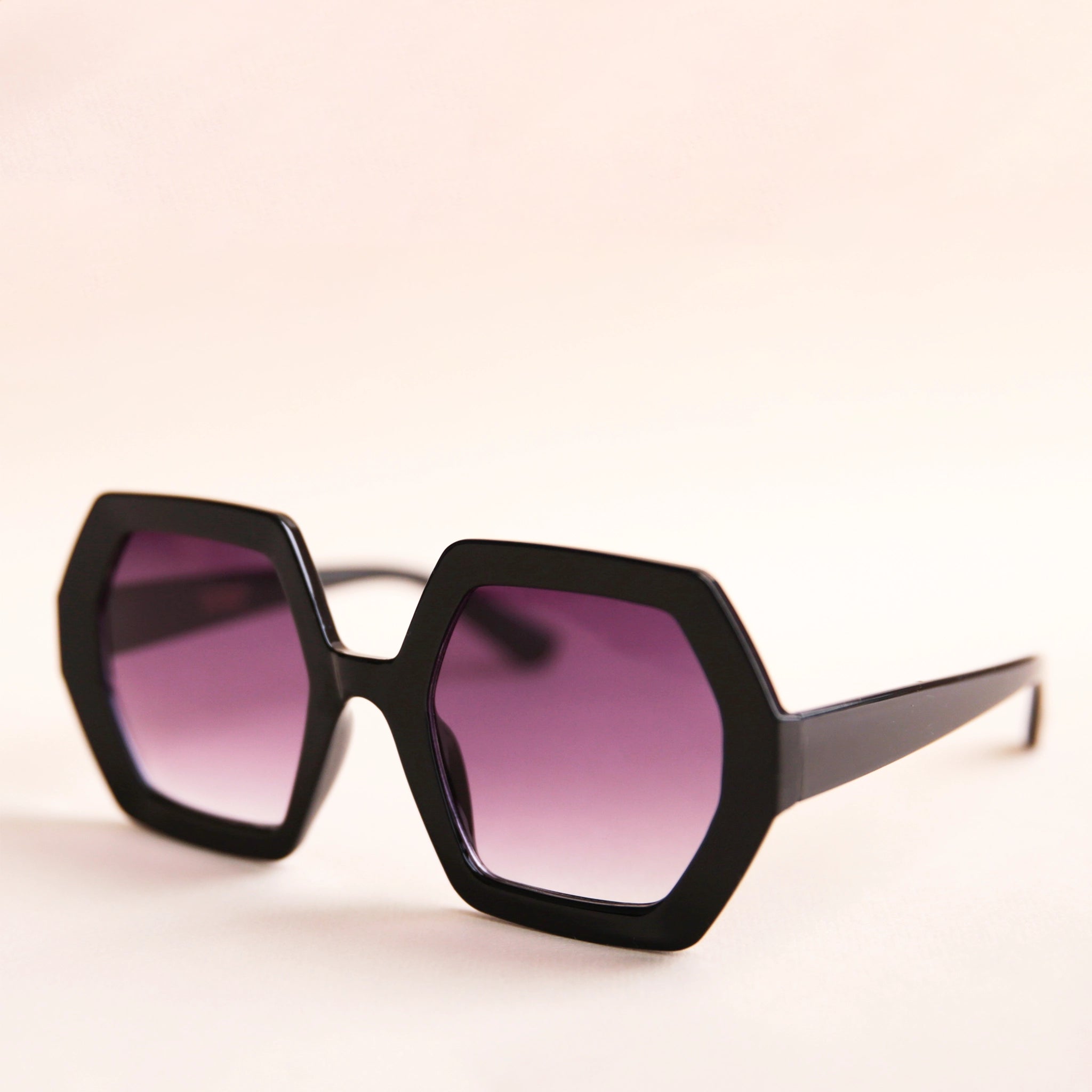 On a neutral background is a pair of hexagon shaped black sunglasses with purple lenses.