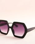 On a neutral background is a pair of hexagon shaped black sunglasses with purple lenses.