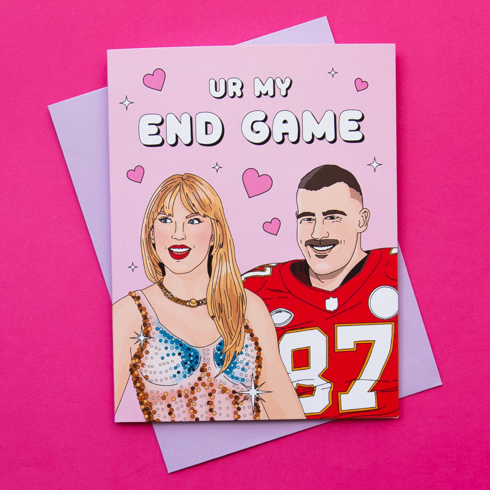 On a hot pink background is a pink card with an illustration that looks like Taylor Swift and Travis Kelce along with text above them that reads, &quot;Ur My End Game&quot;. The card comes with a light purple envelope.