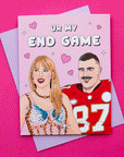 On a hot pink background is a pink card with an illustration that looks like Taylor Swift and Travis Kelce along with text above them that reads, "Ur My End Game". The card comes with a light purple envelope.