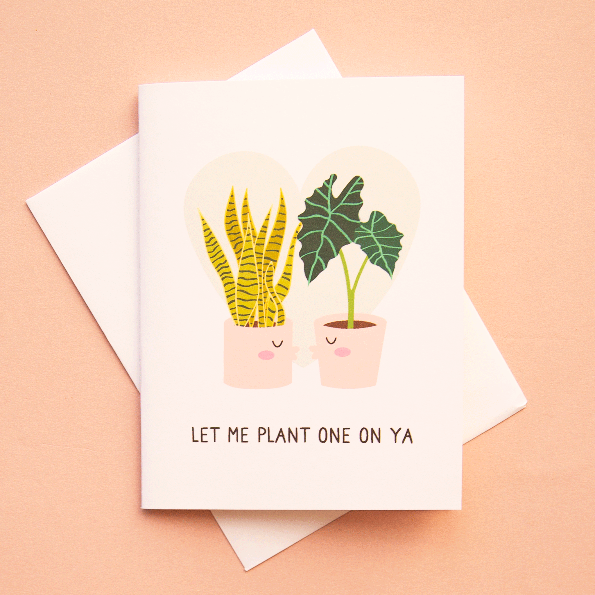  A white card with a light grey heart in the background behind a snake plant and an Alocasia Polly that are planted in two separate pink pots that have kissing faces and are going in for a kiss. There are black words underneath that read, "Let Me Plant One On Ya".
