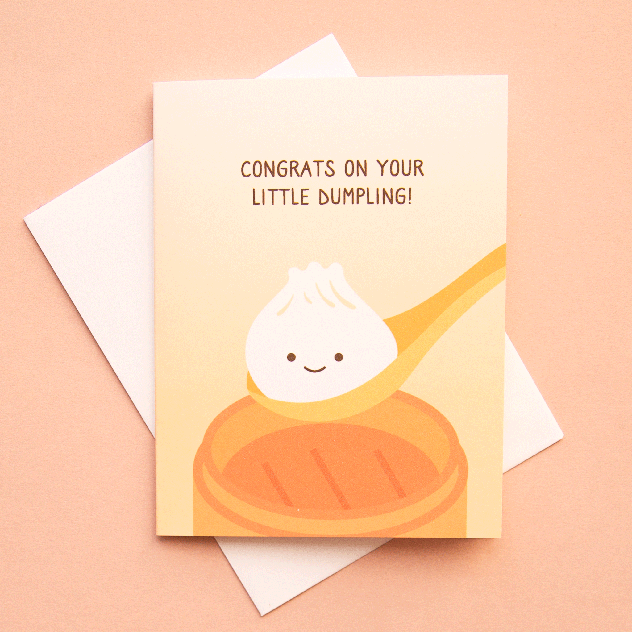 A light orange card with a illustration of a dumpling in a spoon with a smiling face along with text above it that reads, "Congrats On Your Little Dumpling!" in dark brown letters along with a white envelope.