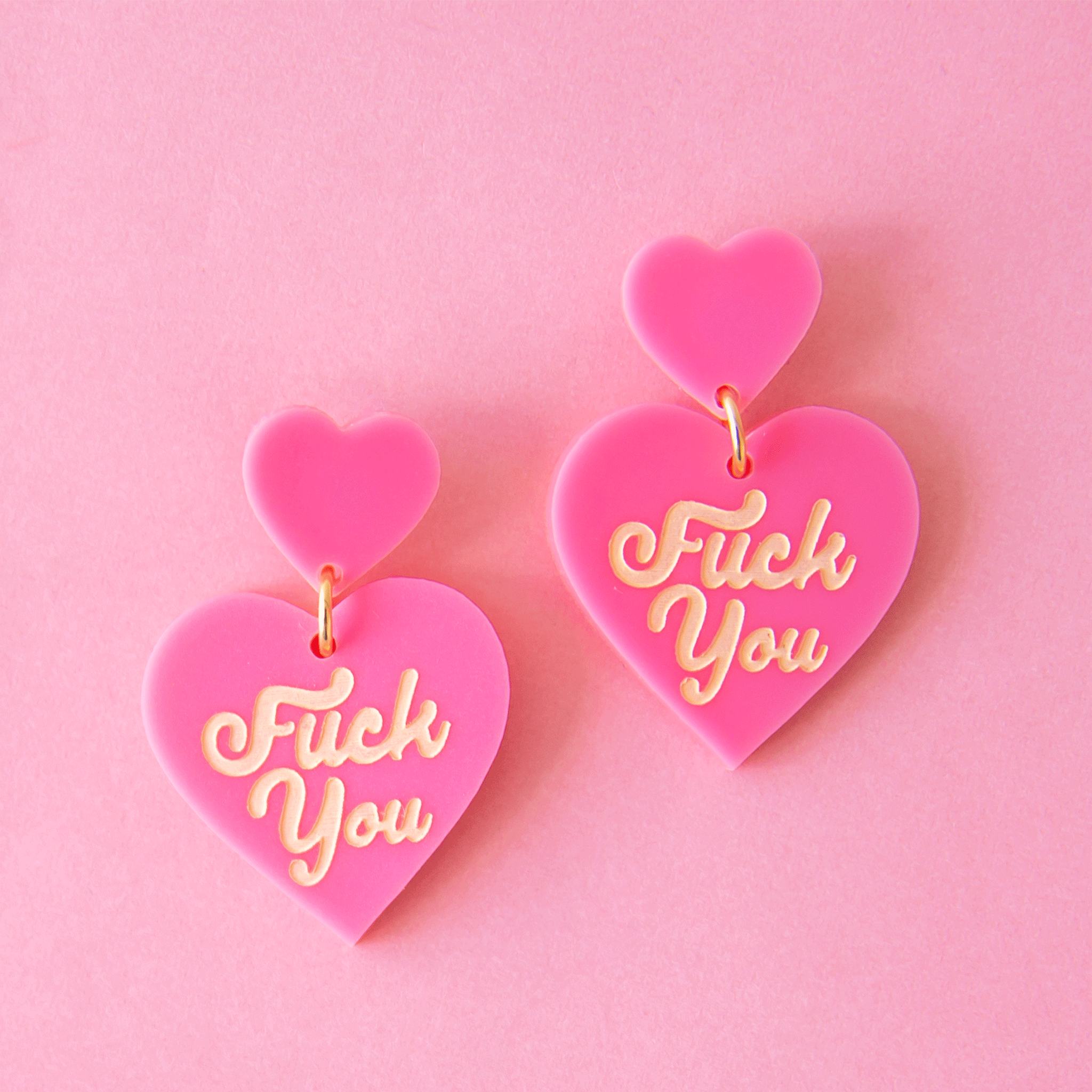On a pink background is a hot pink pair of heart shaped earrings with ivory &quot;Fuck You&quot; on each heart. 