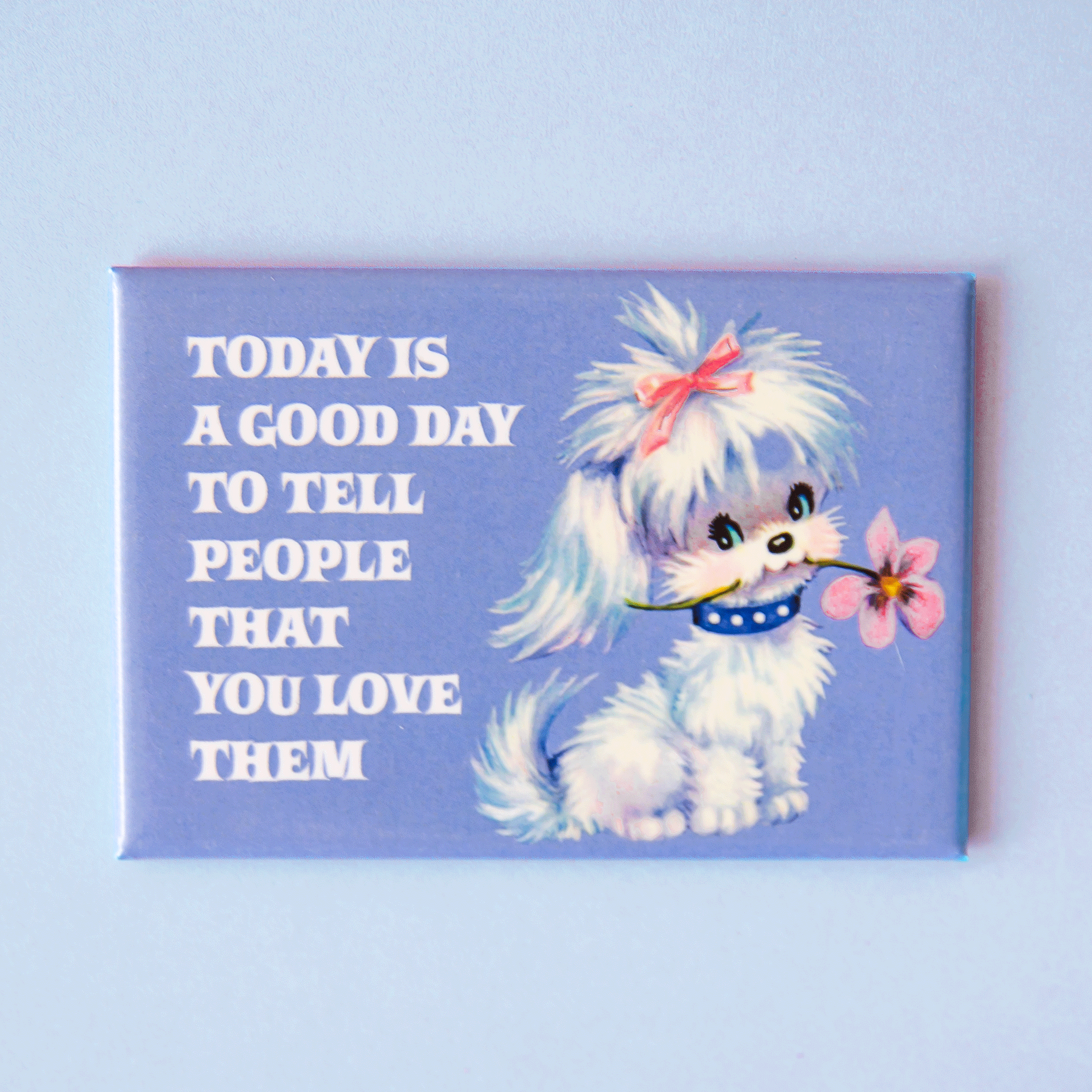 On a blue background is a periwinkle rectangle magnet with a graphic of a white dog holding a flower in its mouth with white text on the left that reads, "Today Is A Good Day To Tell People That You Love Them". 