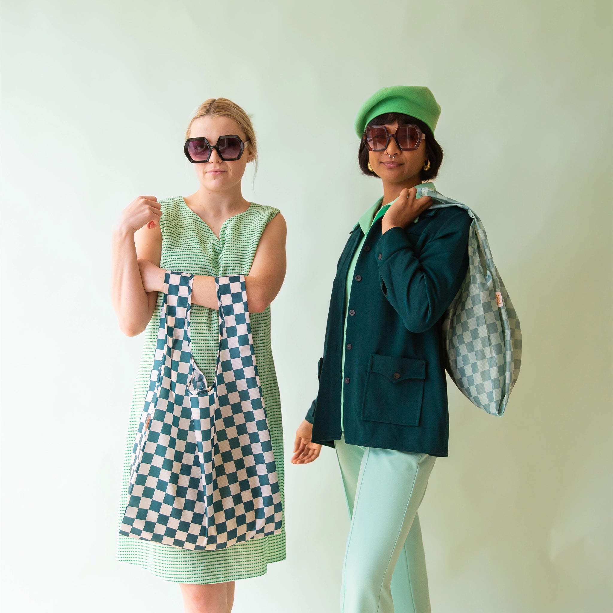 On a light blue background is a model holding a turquoise and light blue checkered print nylon bag next to another model holding our reusable bag in the shade Aquamarine.