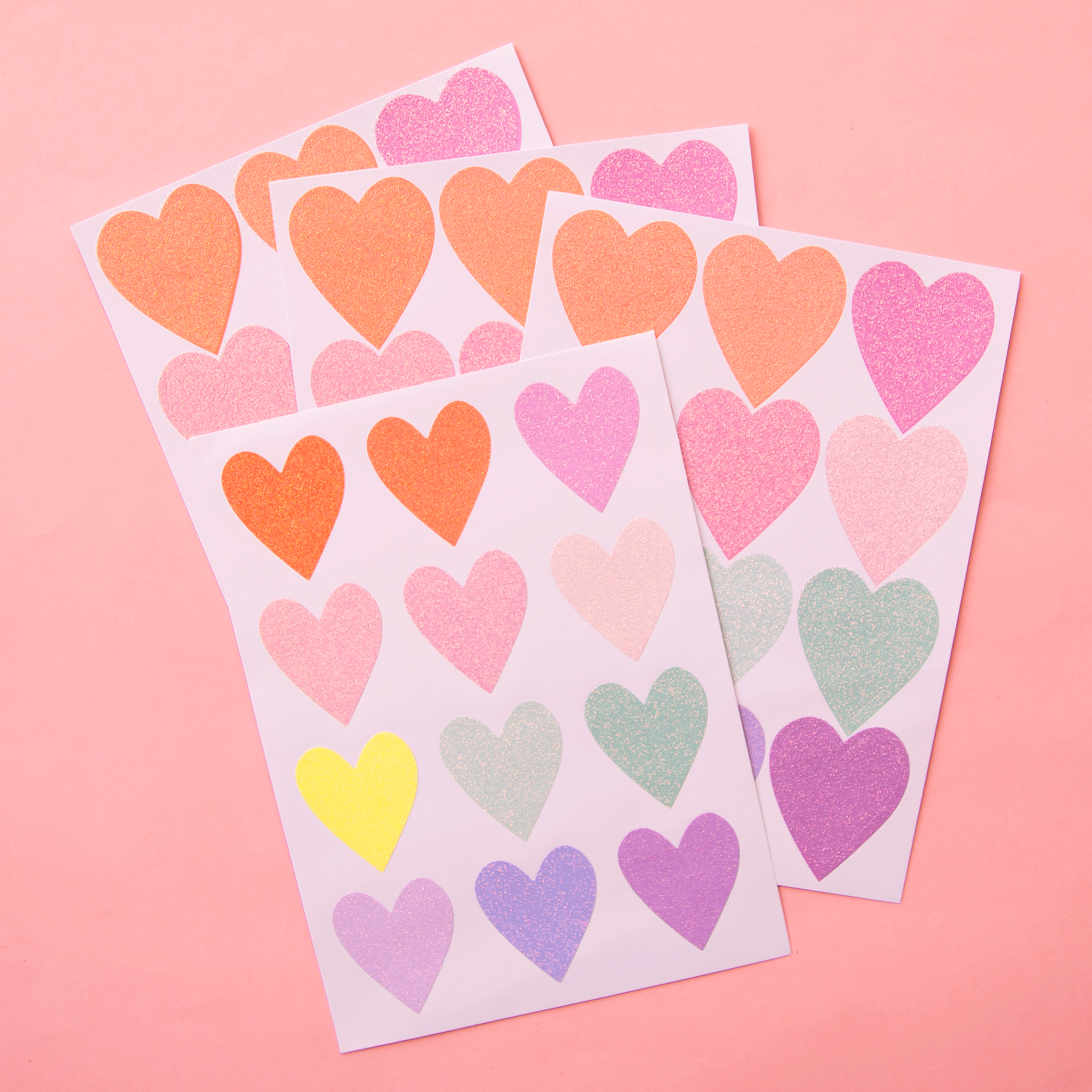 On a pink background is sheets of multi colored heart shaped stickers with glitter on them. 