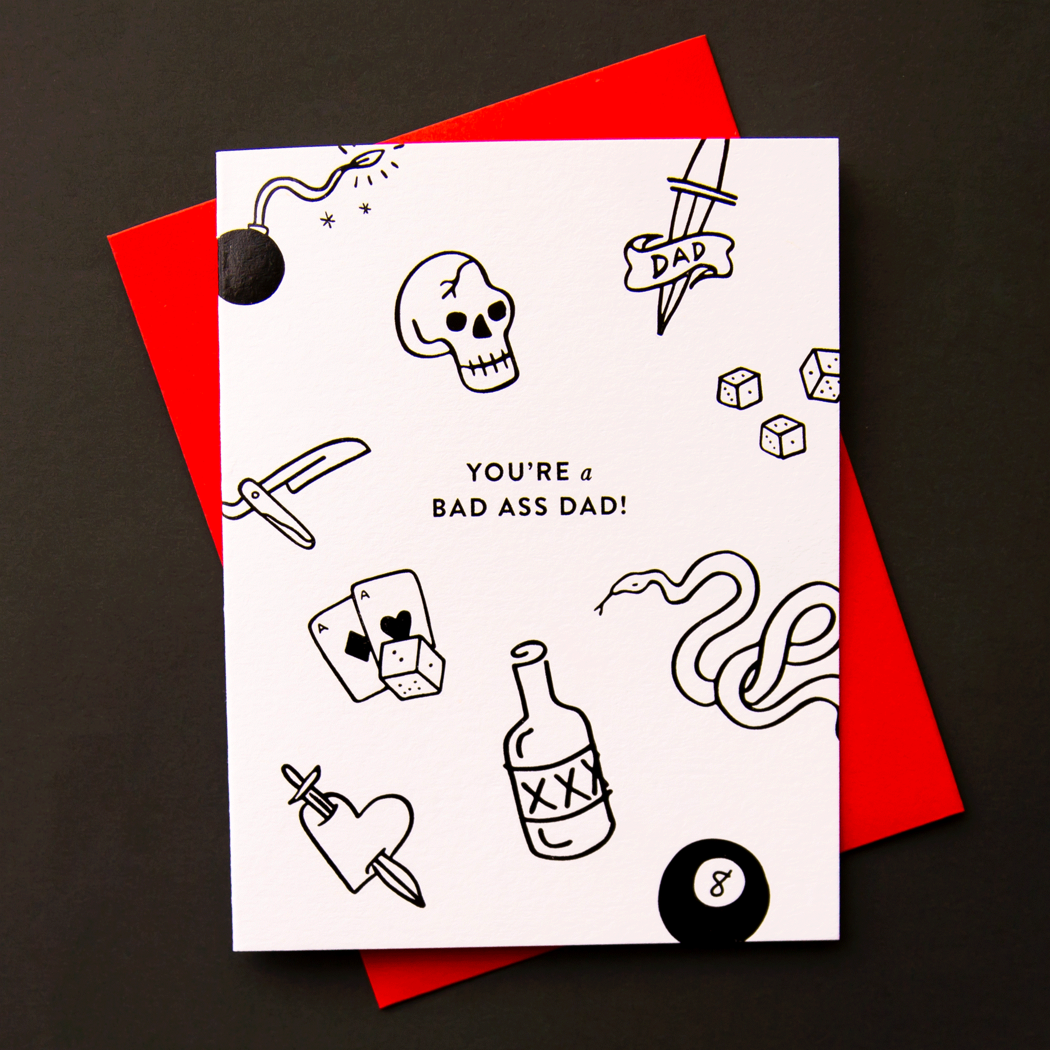 On a black background is a white card with black tattoo-like sketches of skulls, cards, a snake, eight ball and more along with text in the center that reads, "You're a Bad Ass Dad!". 
