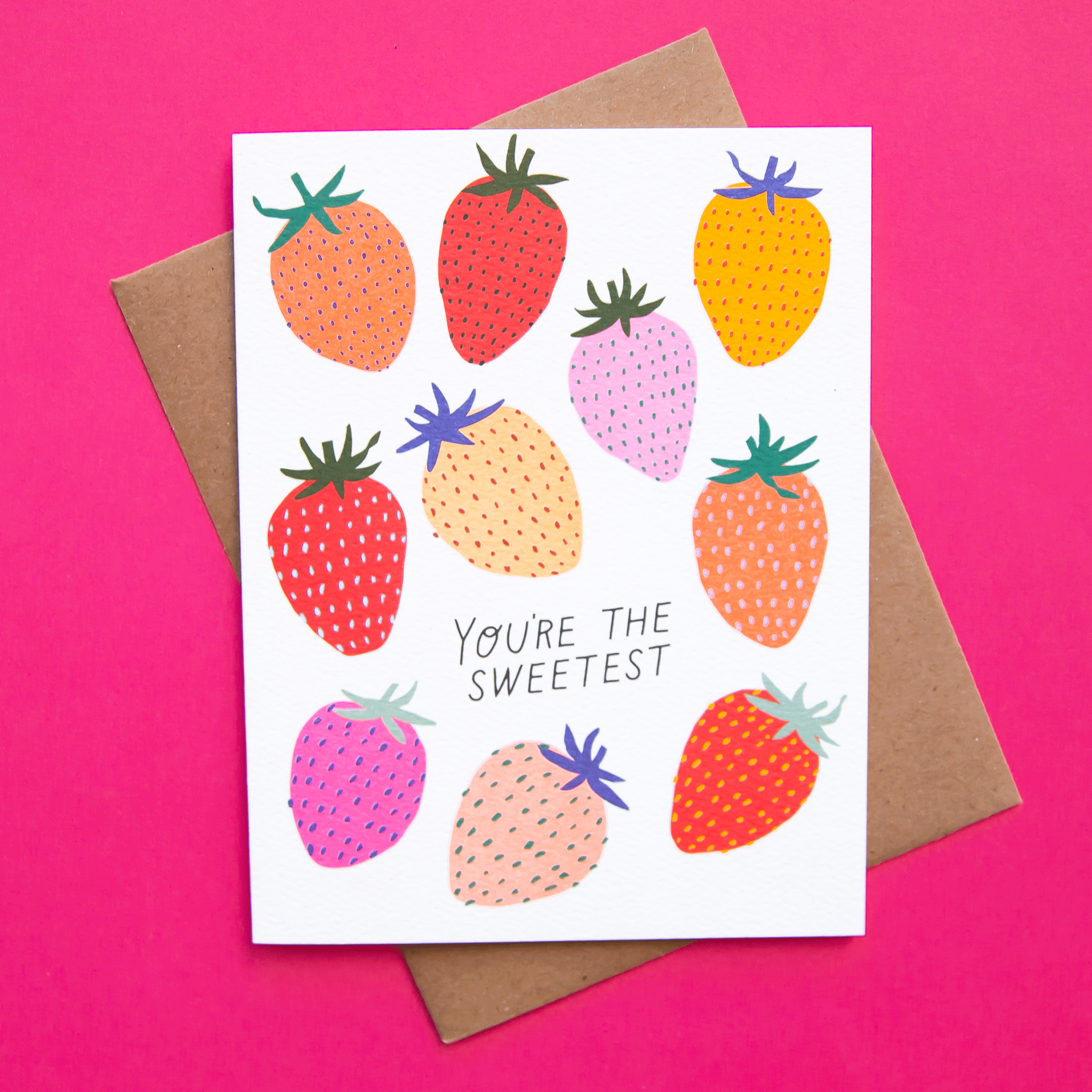 On a hot pink background is a white greeting card with various strawberry prints on the front in shades of red, pink and yellow along with text in the center that reads, "You're The Sweetest".