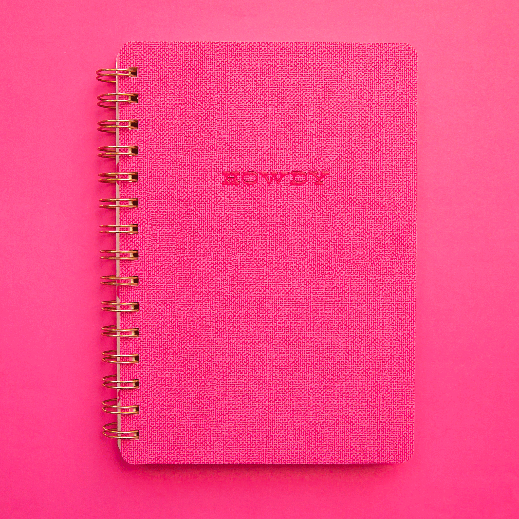 On a pink background is a hot pink spiral bound notebook with a soft cover and text in the center that reads, &quot;Howdy&quot; along with a pink and white horseshoe print on the inside cover. 