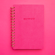 On a pink background is a hot pink spiral bound notebook with a soft cover and text in the center that reads, "Howdy" along with a pink and white horseshoe print on the inside cover. 