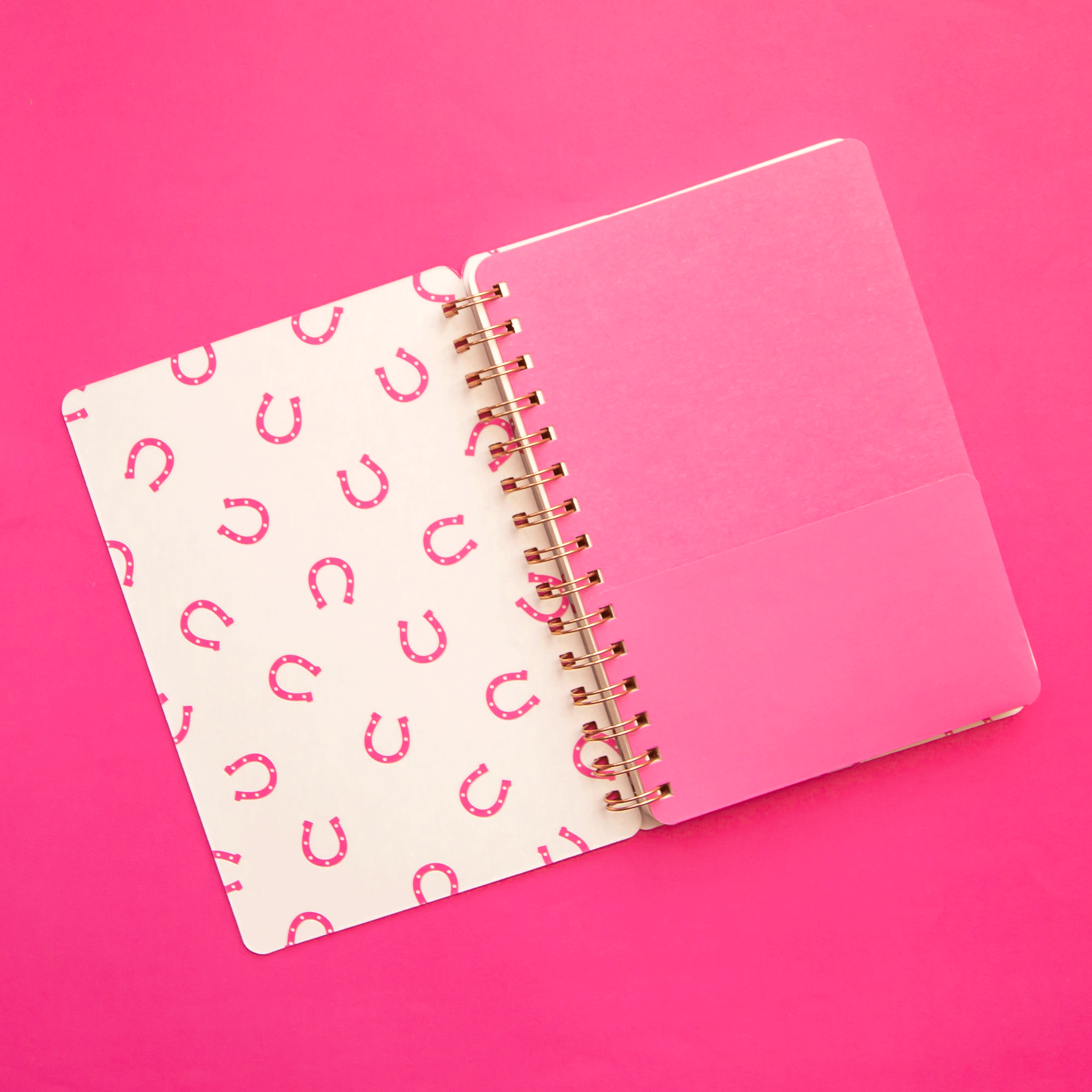 On a pink background is a hot pink spiral bound notebook with a soft cover and text in the center that reads, "Howdy" along with a pink and white horseshoe print on the inside cover.