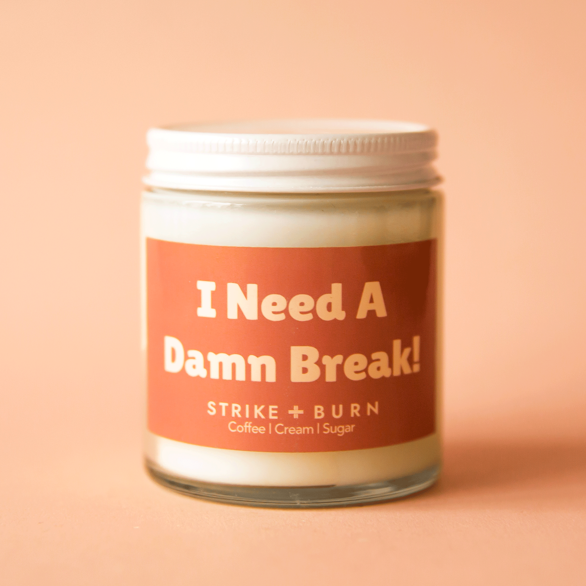 On a peach background is a clear glass jar candle with a warm brown label that reads, &quot;I Need A Damn Break!&quot;. 
