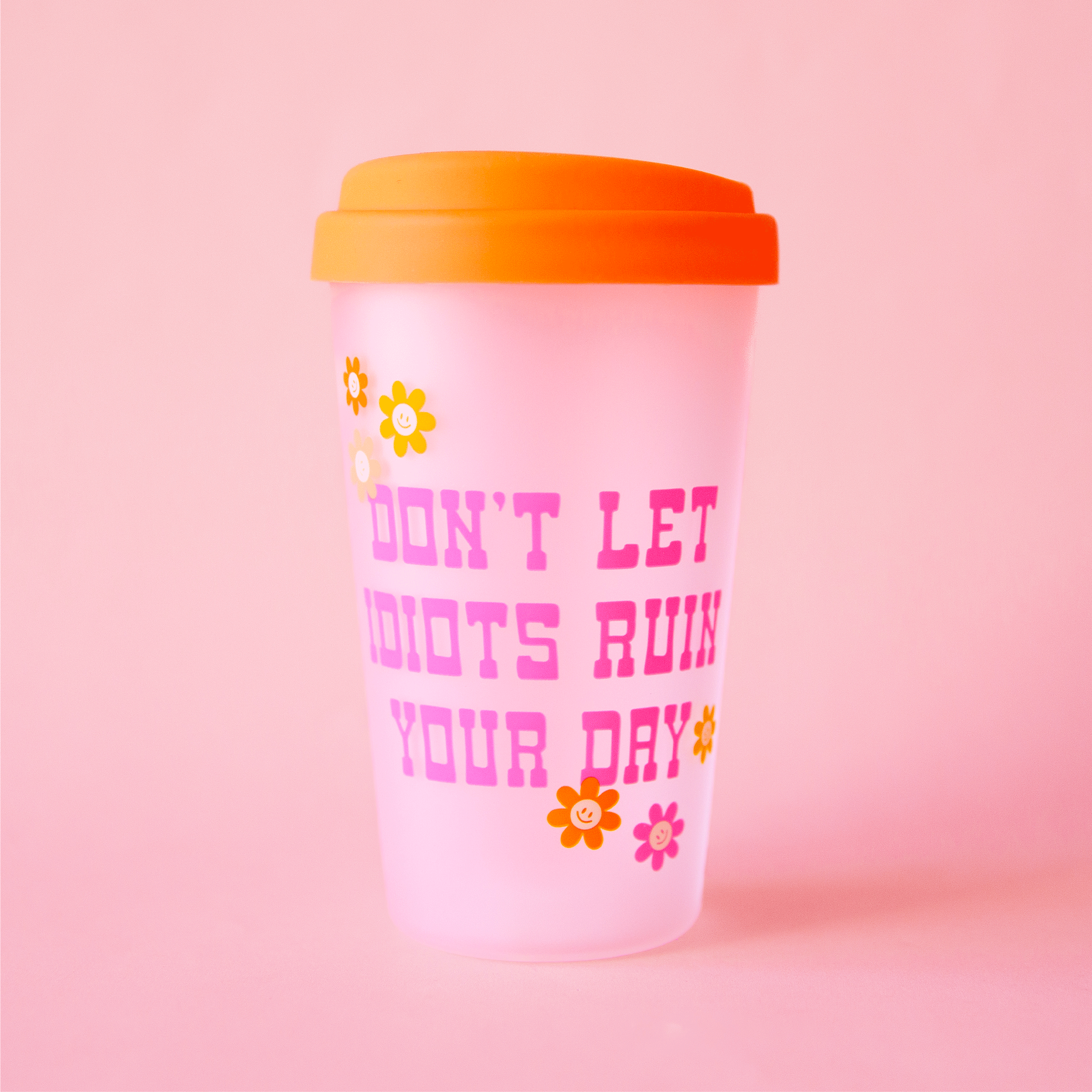 On a pink background is a clear tumbler with a orange silicone lid and pink text on the front that reads, &quot;Don&#39;t Let Idiots Ruin Your Day&quot;. 