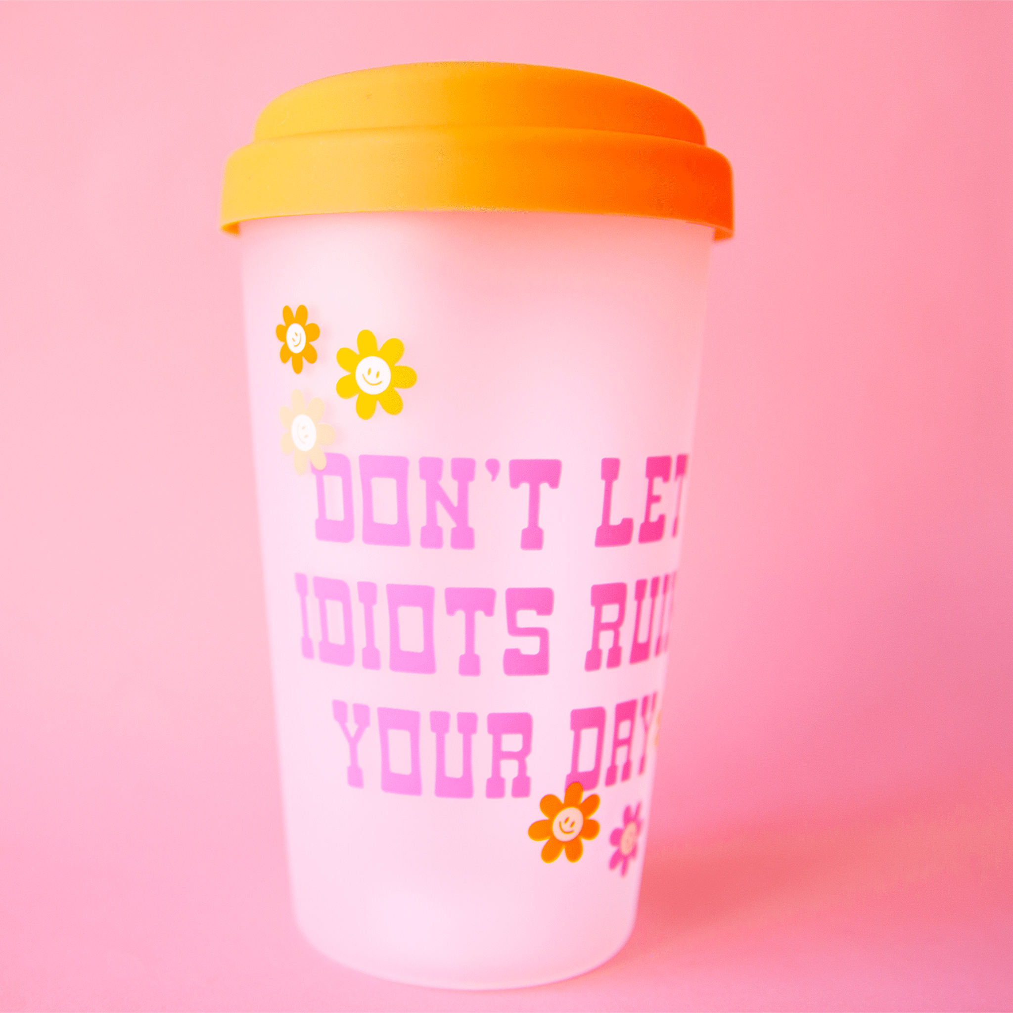 On a pink background is a clear tumbler with a orange silicone lid and pink text on the front that reads, &quot;Don&#39;t Let Idiots Ruin Your Day&quot;.