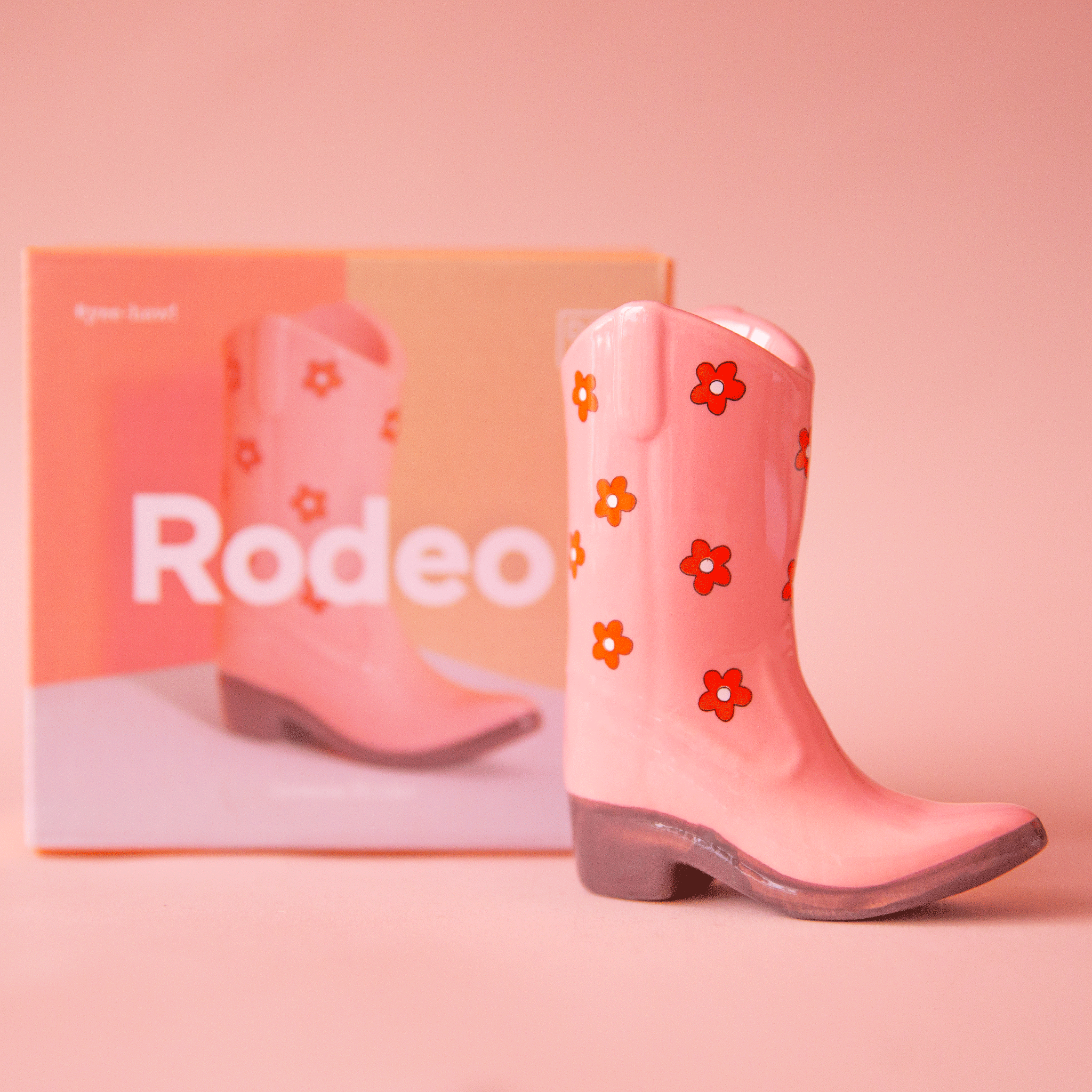On a peach background is a pink ceramic cowboy boot shaped incense holder with a red flower print. 