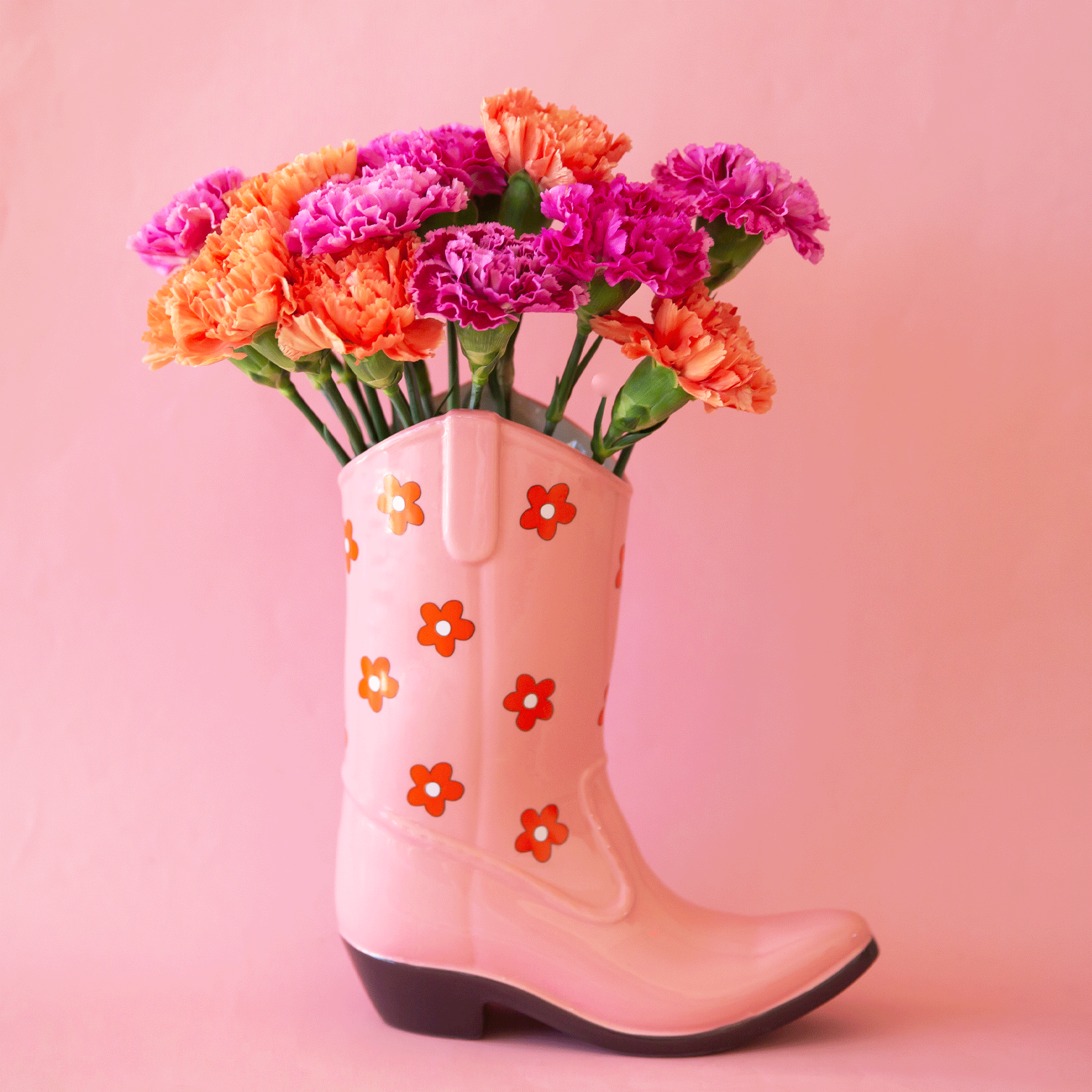 On a pink background is a pink cowboy boot shaped vase with a red and white daisy print on it. Flowers pictured not included with purchase.
