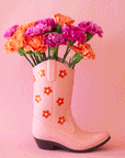 On a pink background is a pink cowboy boot shaped vase with a red and white daisy print on it. Flowers pictured not included with purchase.