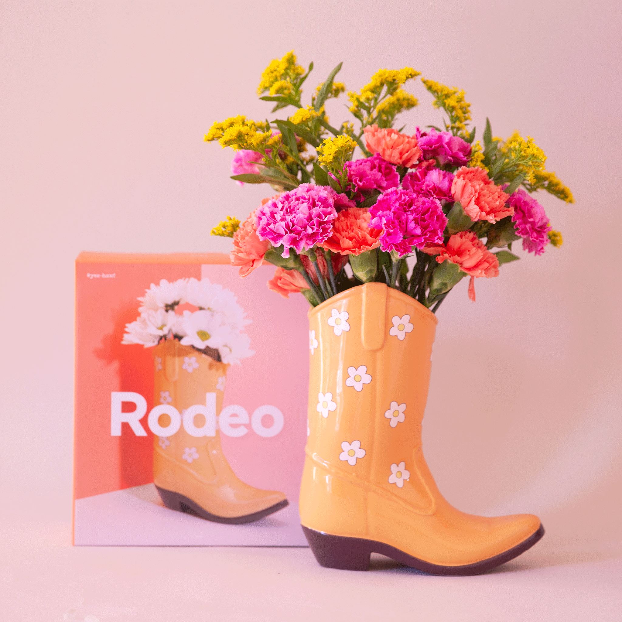 On a pink background is a yellow cowboy boot shaped vase with white and yellow daisy print all over. Flowers not included with purchase