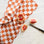 On a marble backdrop is an orange and ivory checkered kitchen towel with a staged blood orange cut up next to it. 
