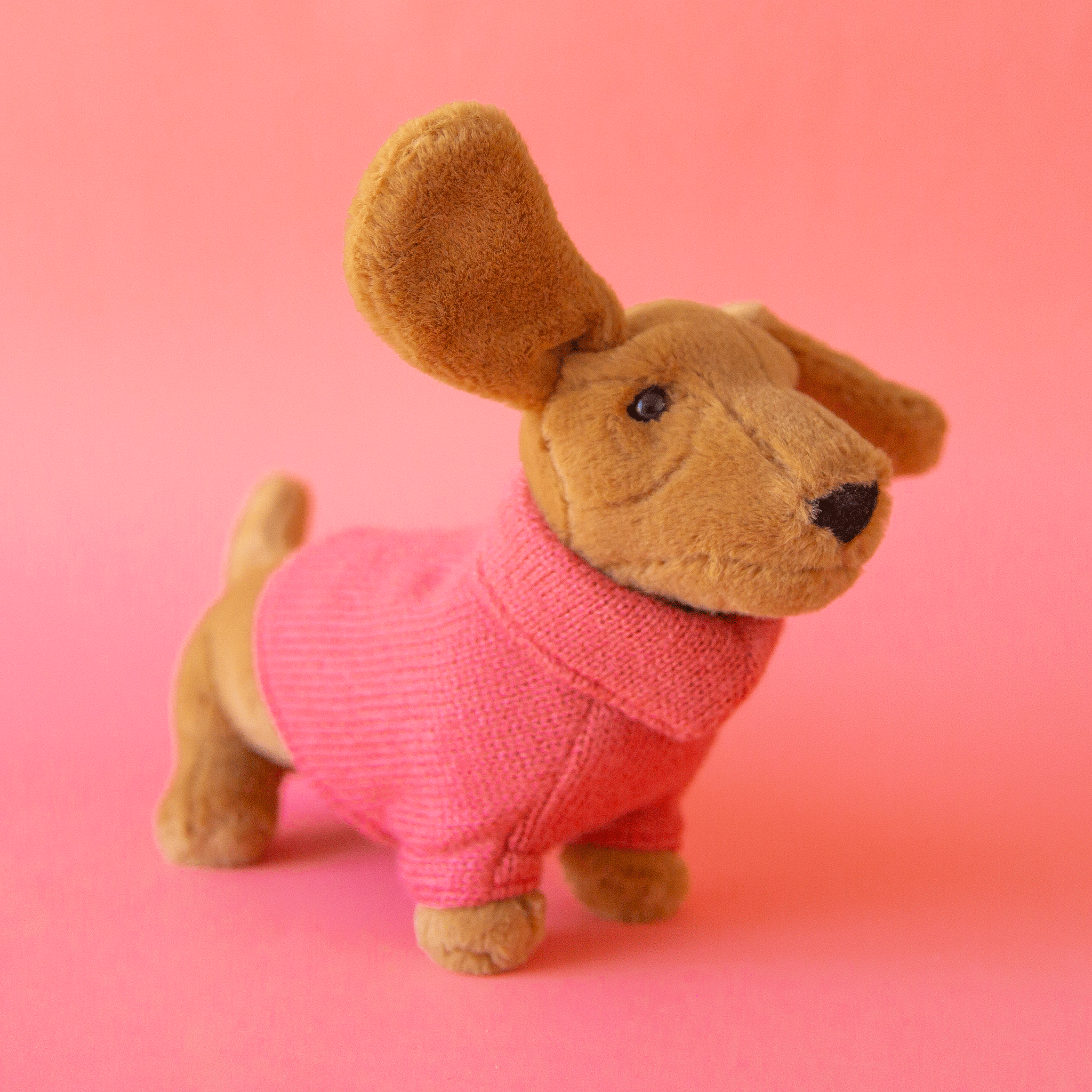On a pink background is a tan sausage dog stuffed animal wearing a pink sweater.