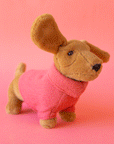 On a pink background is a tan sausage dog stuffed animal wearing a pink sweater.