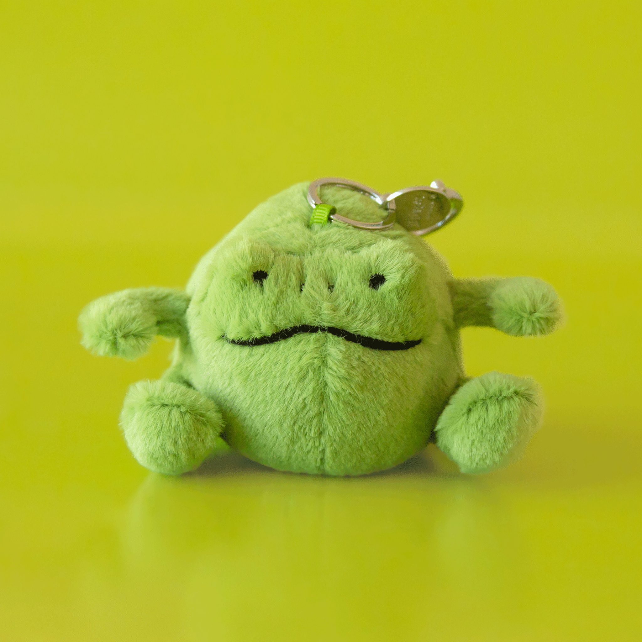 On a green background is a small green stuffed toy frog shaped bag charm.